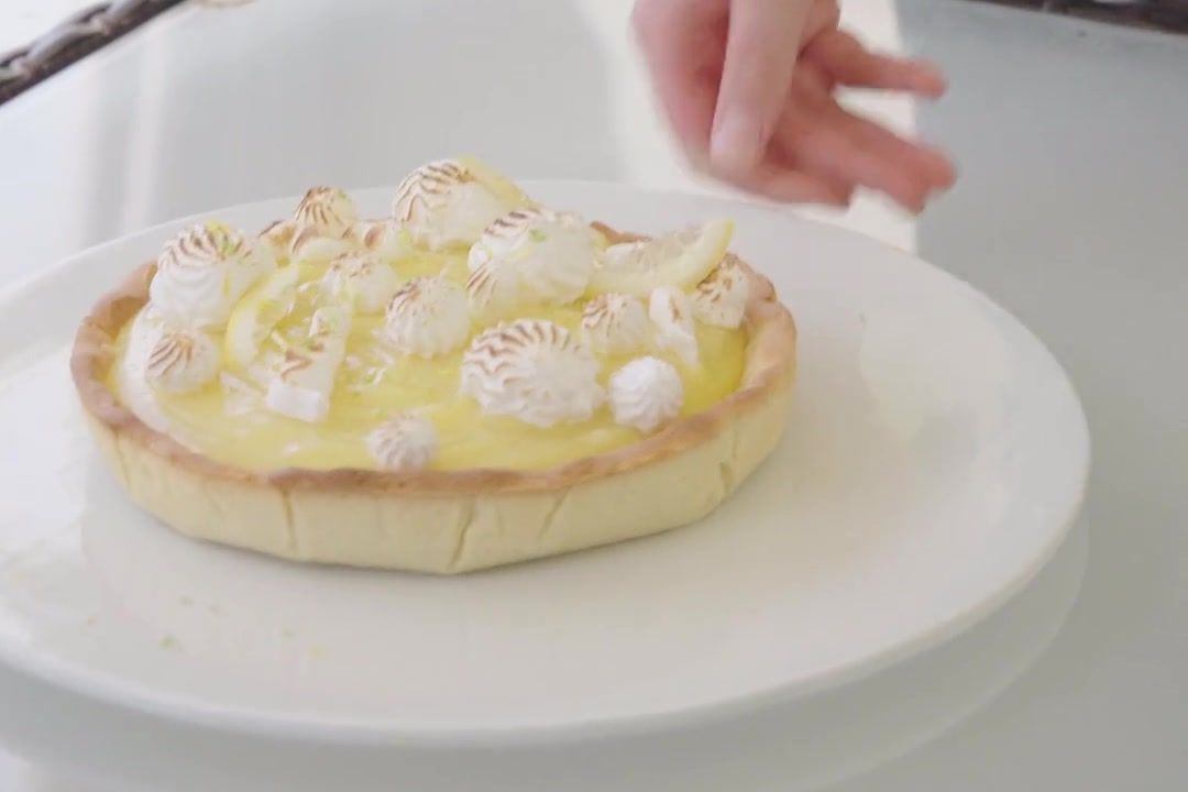 Une tarte au citron meringuée posée sur une assiette blanche, dehors sur une table en verre, avec ses meringues dorées au chalumeau